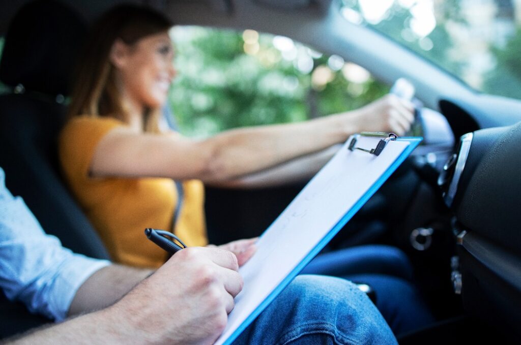 Nahaufnahme eines Fahrlehrers, der eine Checkliste in der Hand hält, während im Hintergrund eine Fahrschülerin das Auto lenkt und fährt. Erwerb des Führerscheins.