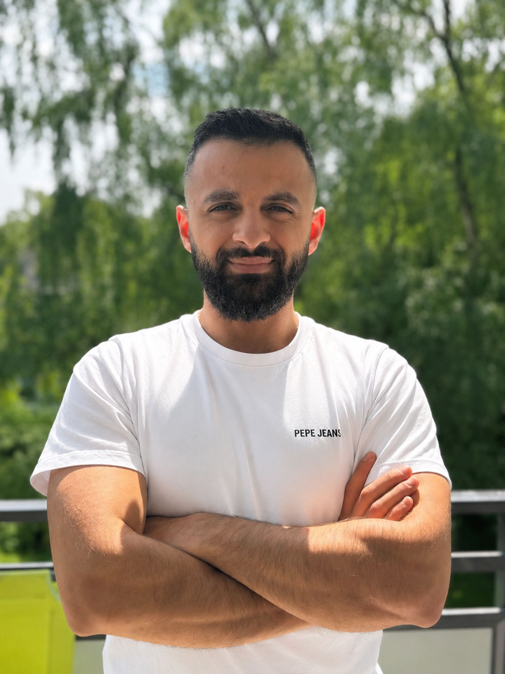Ein Mann mit dunklem Haar trägt ein weißes T Shirt und verschränkt die Arme draußen