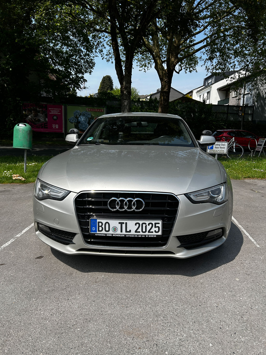 Silbergrauer Audi steht auf einem Parkplatz vor Bäumen und Häusern