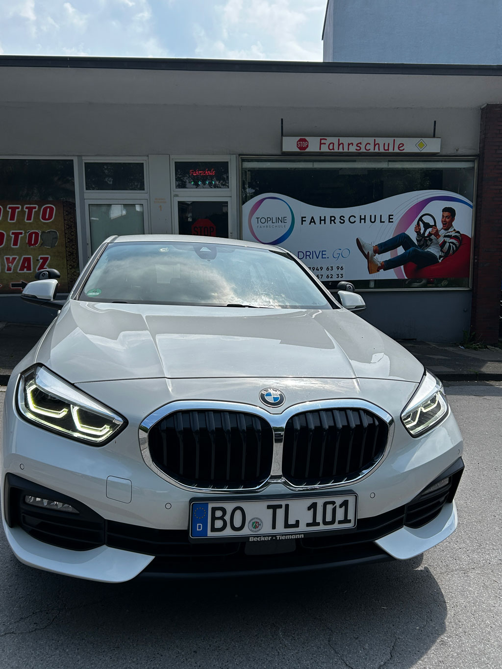 Ein weißer BMW mit leuchtenden Scheinwerfern steht vor einer Fahrschule
